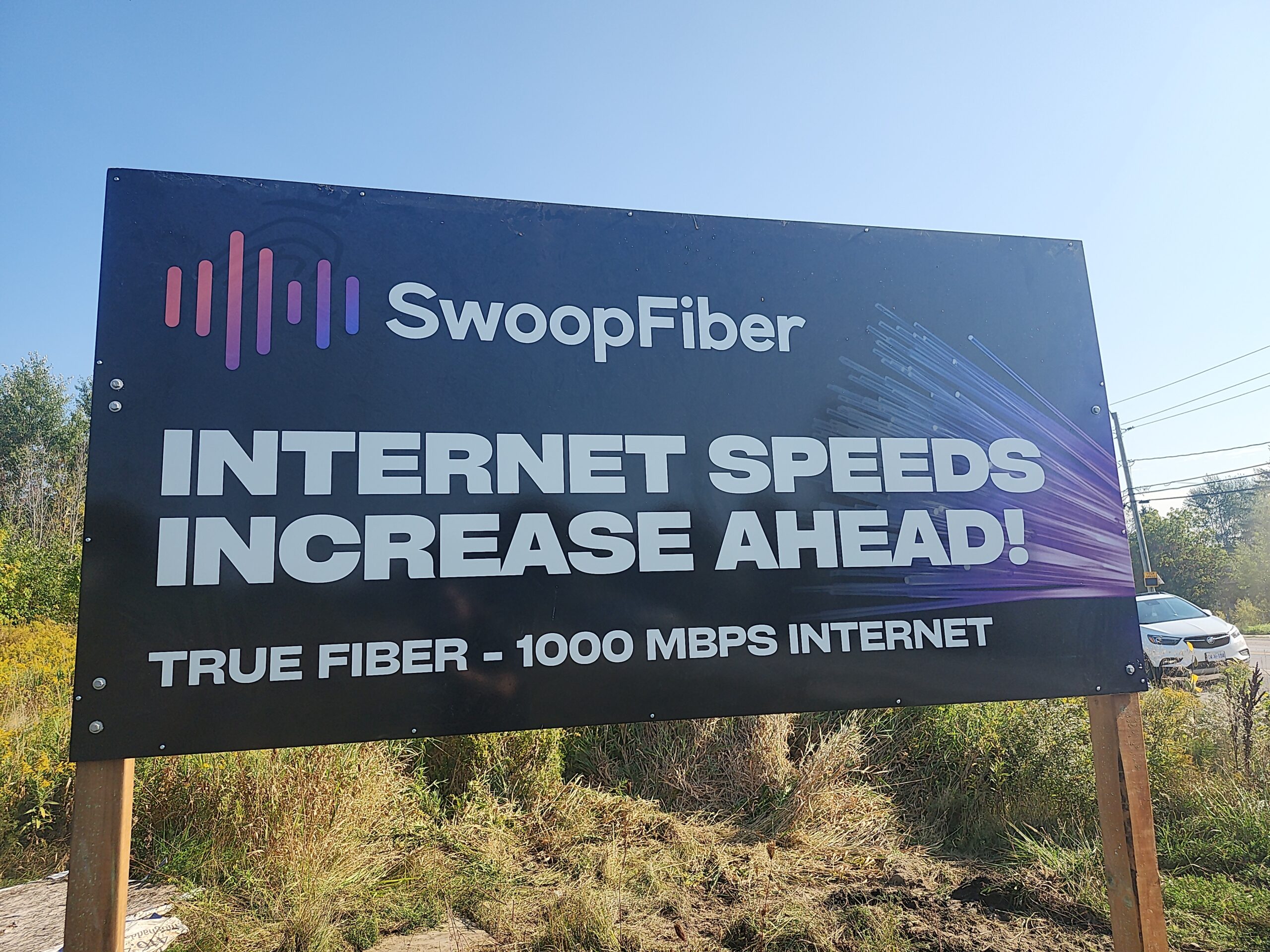 Billboard in a rural area displaying “Internet Speeds Increase Ahead” for SwoopFiber, mounted on posts and surrounded by grass, trees, and open countryside under sunlight.